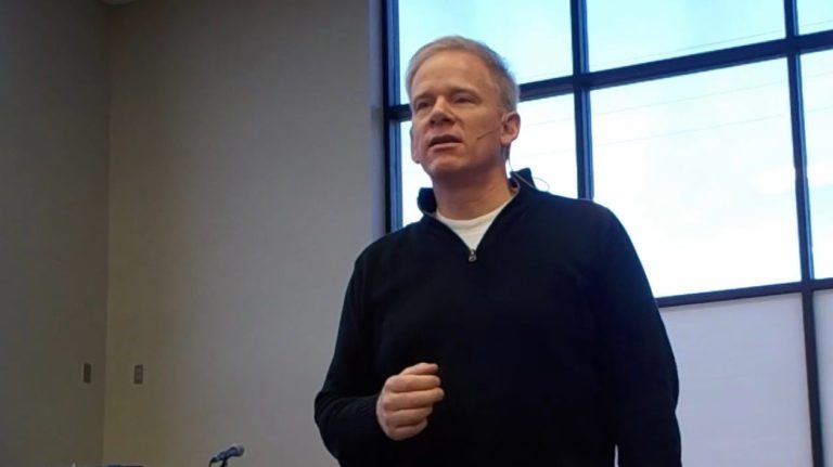 A man is standing and speaking with a calm expression